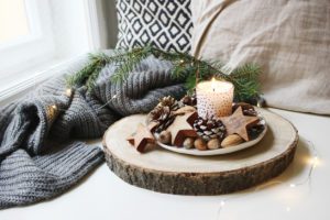 Winter festive still life scene. Burning candle decorated by wooden stars, hazelnuts and pine cones standing near window on wooden cut board. 